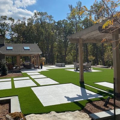 A modern backyard patio with large white pavers set in artificial green turf, a pergola, and a covered lounge area.