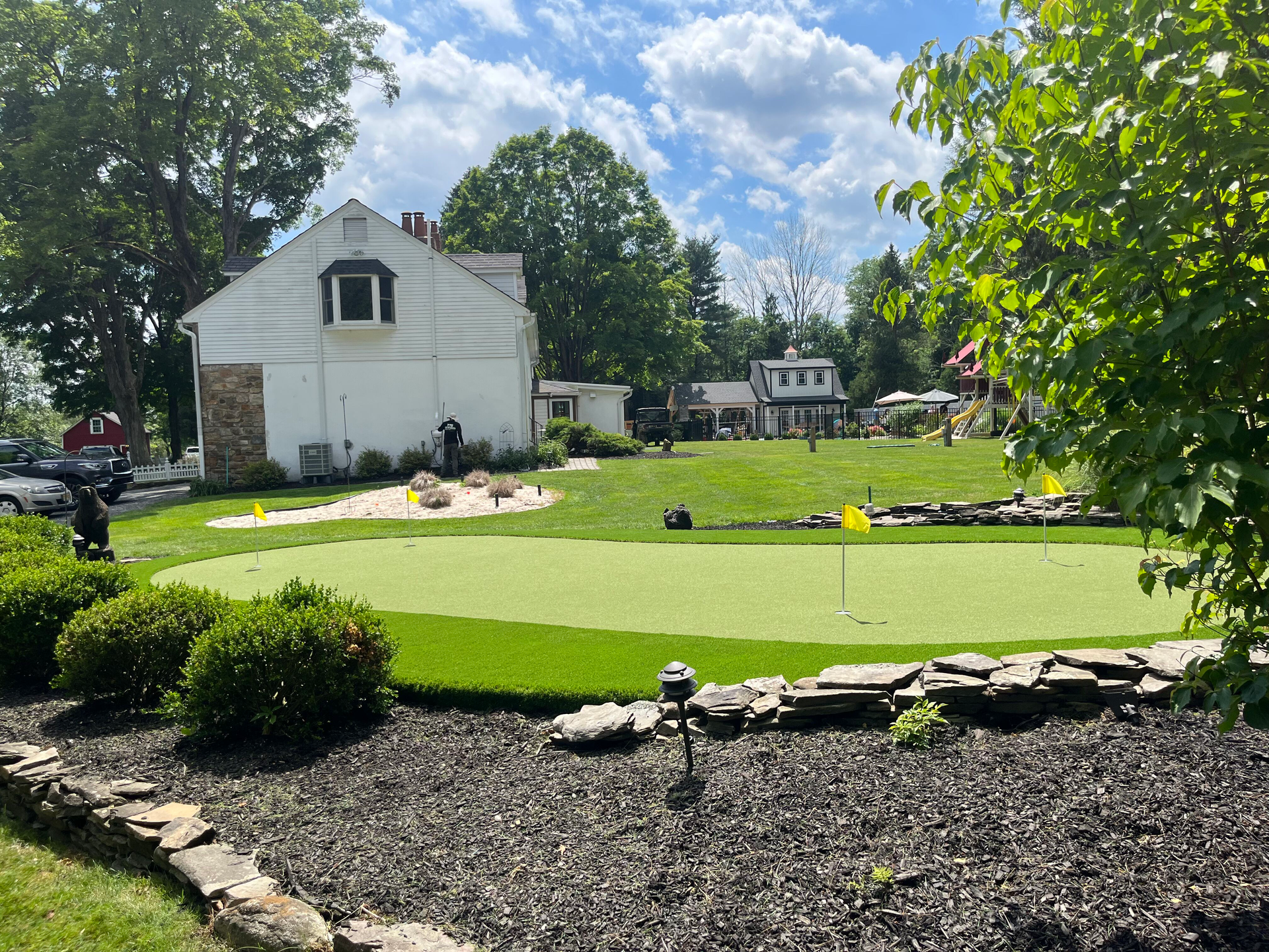 A putting green with several holes and yellow flags sits in a backyard near a white house, surrounded by landscaping.