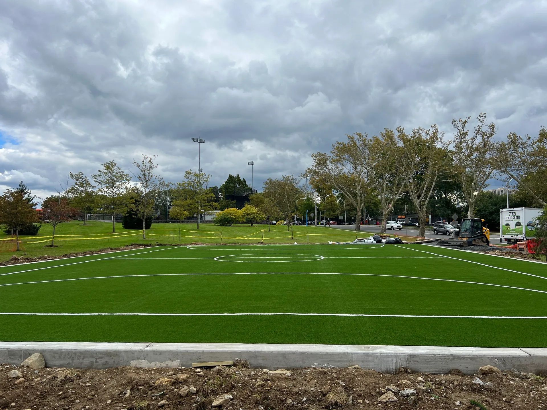 A small, rectangular artificial turf sports field with white markings, bordered by concrete, under a cloudy sky.