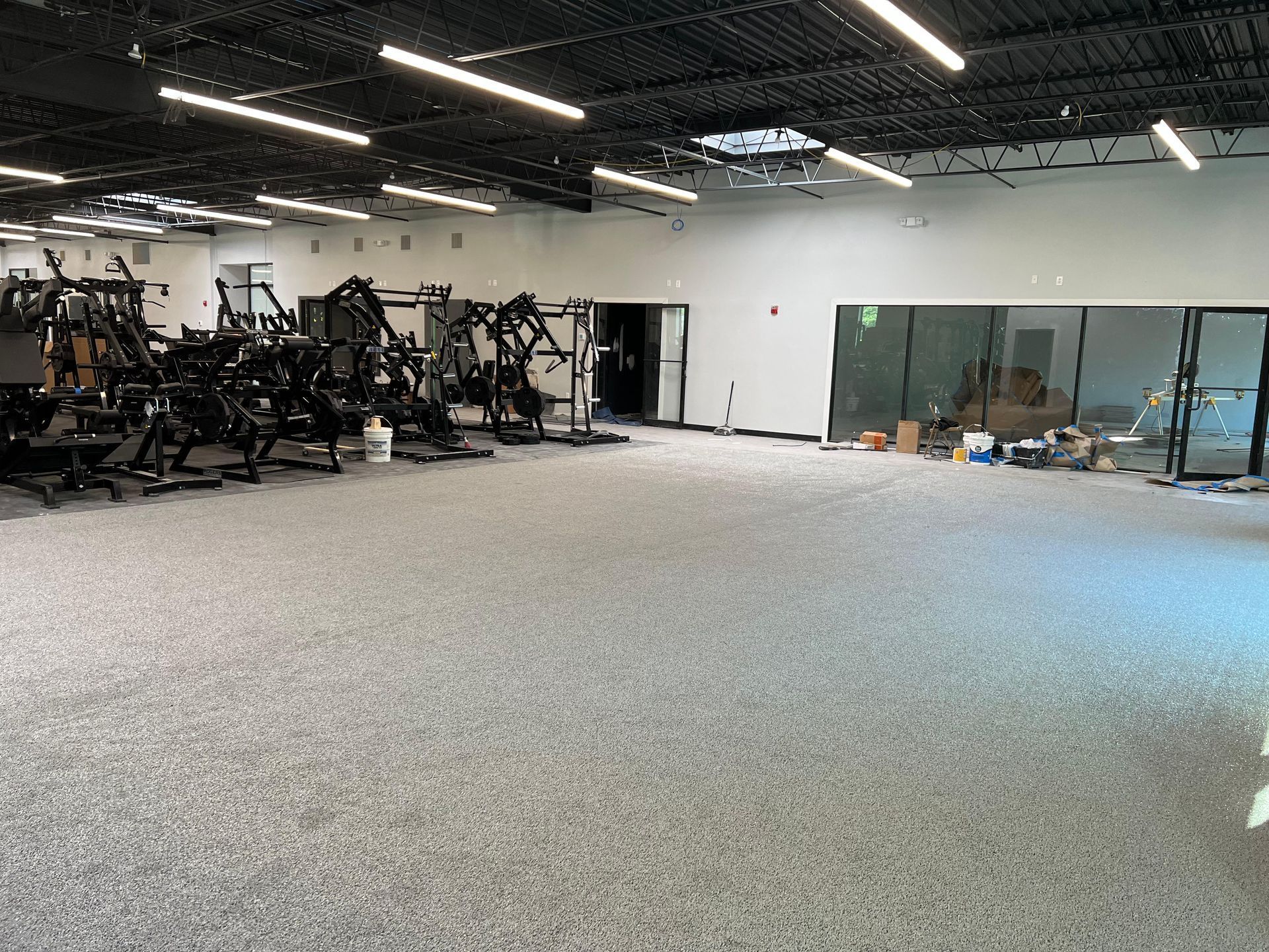 A wide-angle view of a gym interior with exercise equipment on the left, grey speckled flooring, and an open layout.