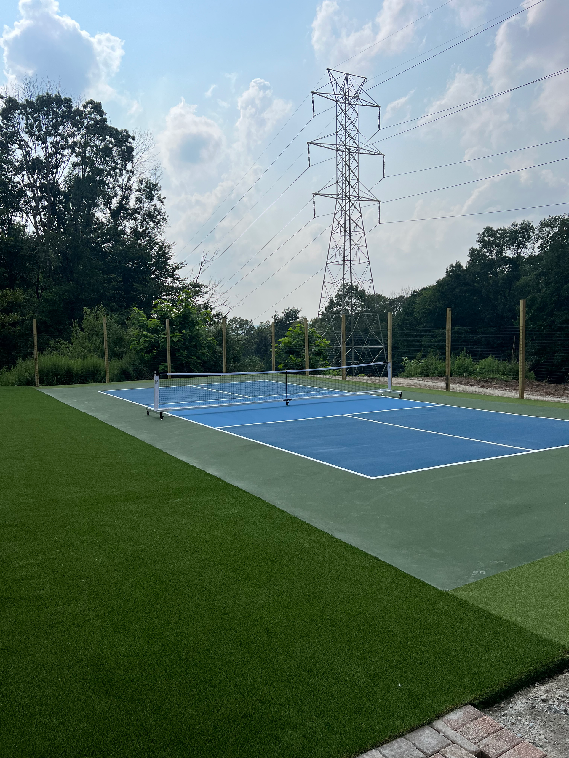 A blue and green pickleball court outdoors, surrounded by a fence, grass, and a large electrical tower under a sunny sky.