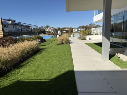 Modern building patio with a tiled walkway, manicured lawn, and ornamental grasses beside a swimming pool under a blue sky.