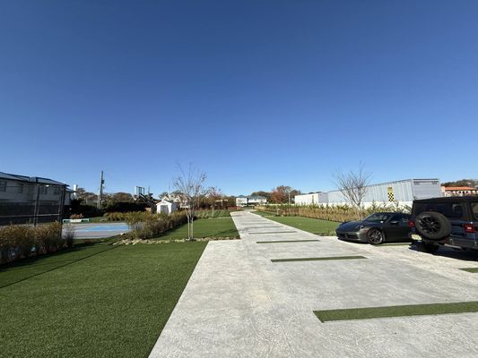 A paved parking area with green strips between spaces, bordered by lawns, a building, and parked cars under a clear sky.