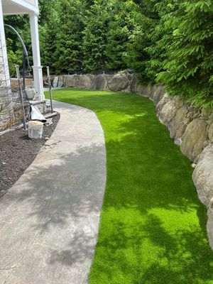 A stone walkway curves alongside a vibrant green lawn bordered by a stacked stone retaining wall and lush trees.