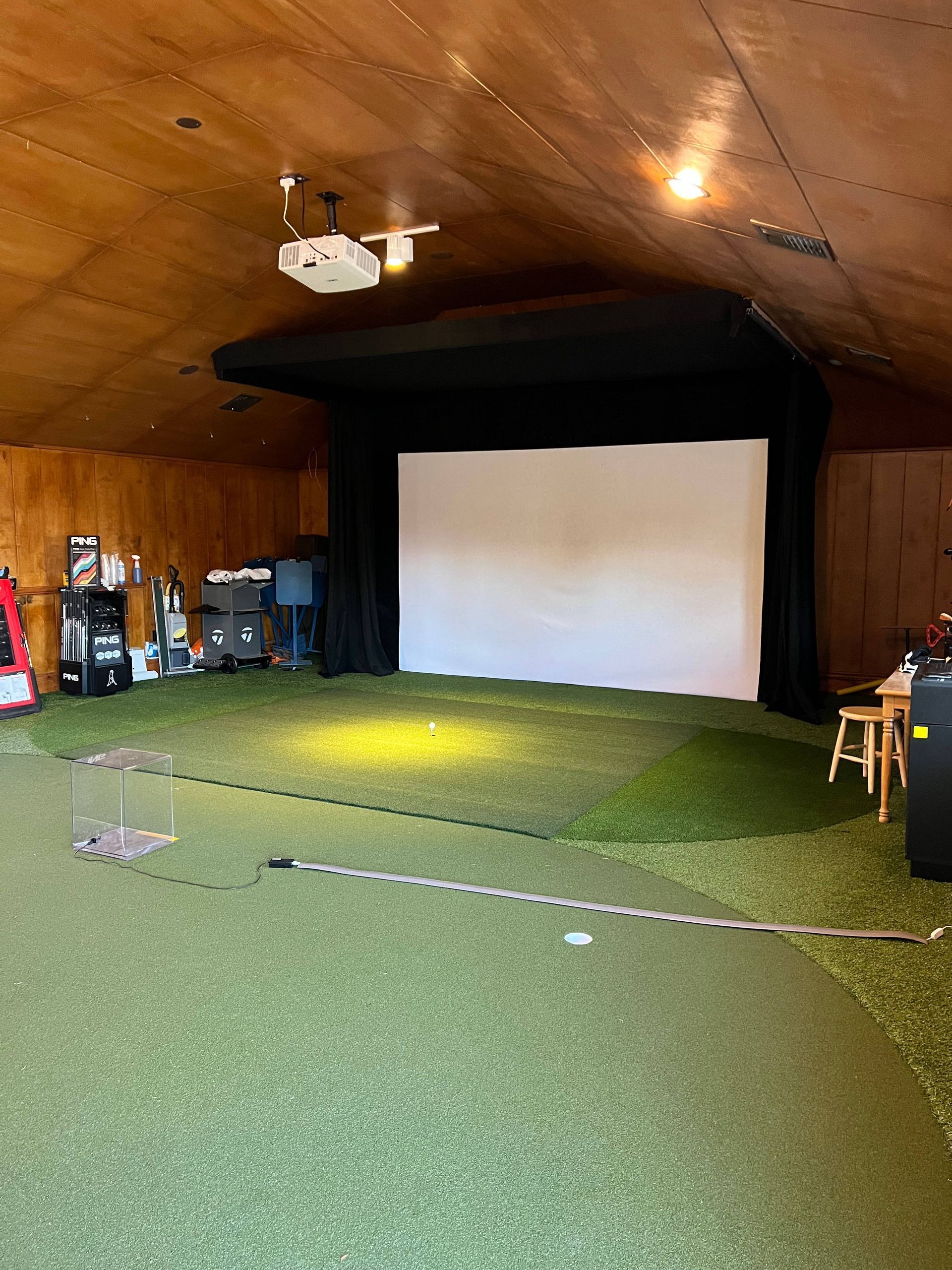 An indoor golf simulator setup featuring a white projection screen, turf floor, and ceiling-mounted projector.