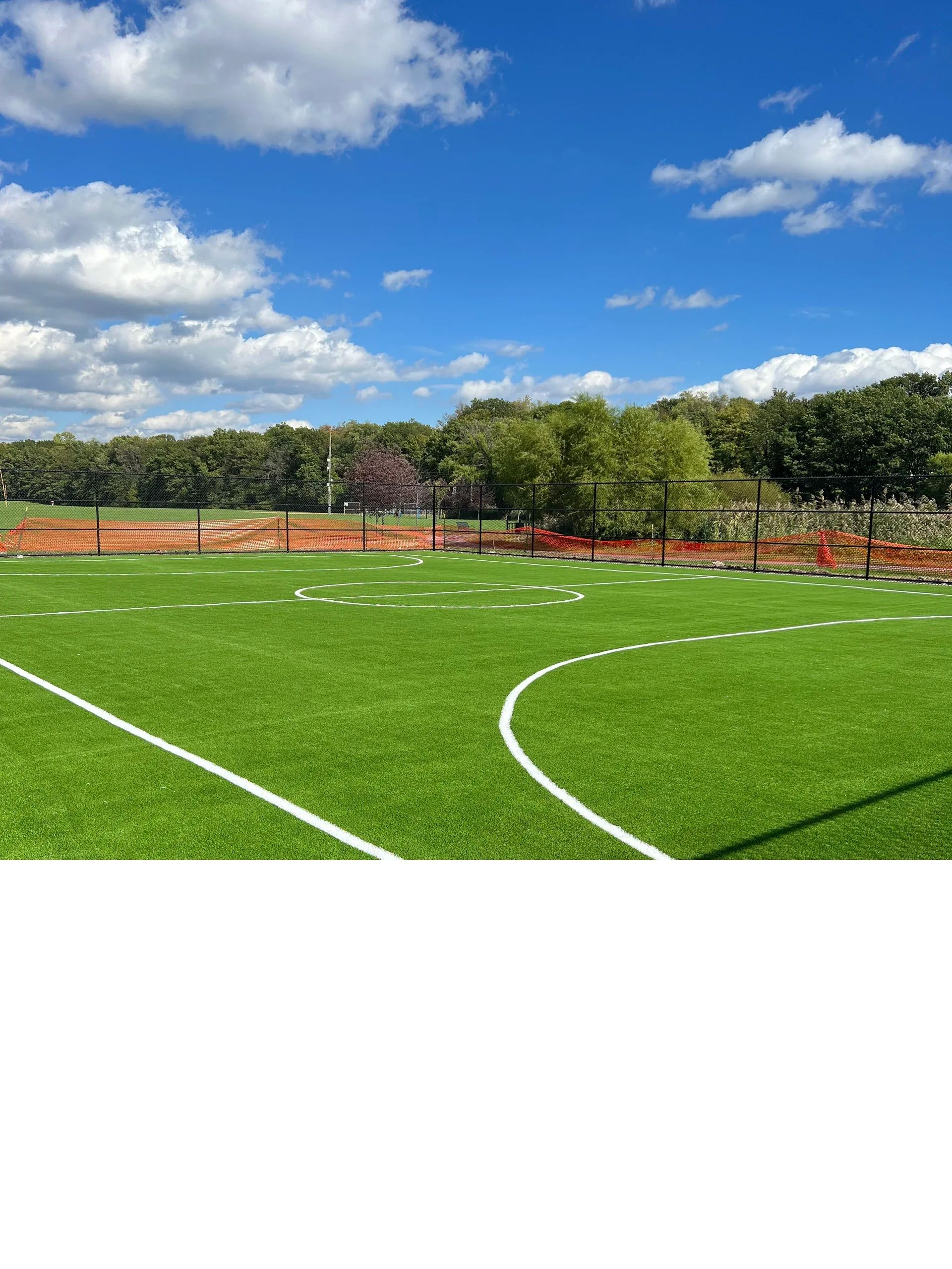 A bright green soccer field with white markings under a vibrant blue sky with scattered clouds and a distant treeline.