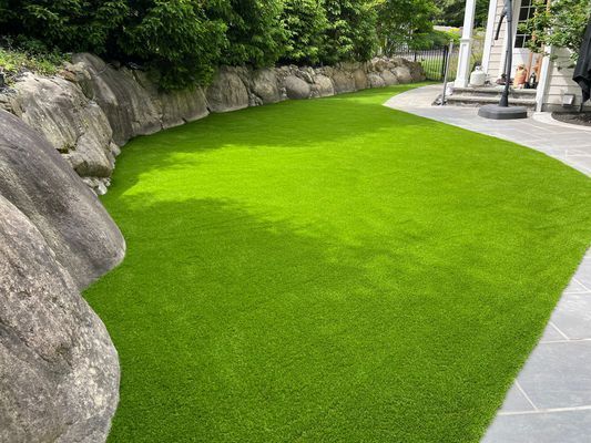 A vibrant, neatly trimmed patch of artificial green grass curved next to large, textured gray rocks and a stone patio.