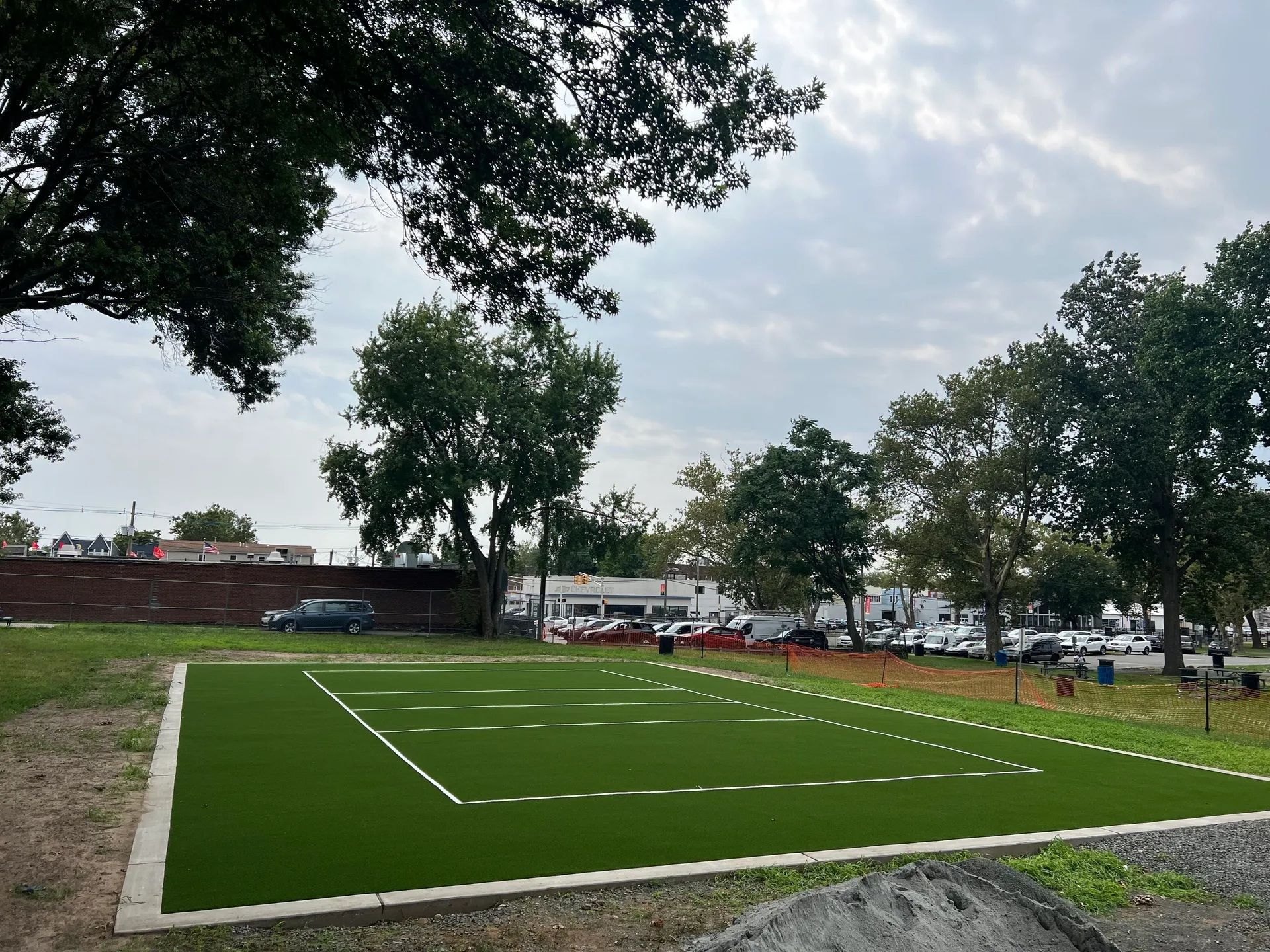 A rectangular artificial turf field with white boundary lines sits in a grassy, tree-lined lot next to a parking area.