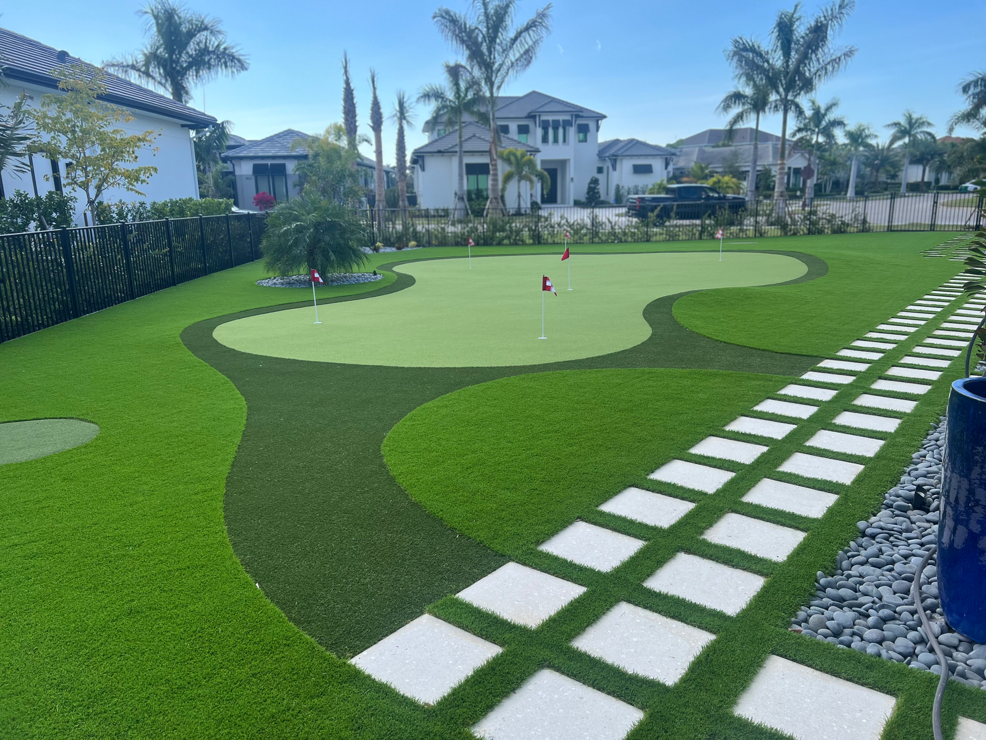 A backyard putting green with artificial grass, featuring a stone paver walkway and palm trees under a clear blue sky.