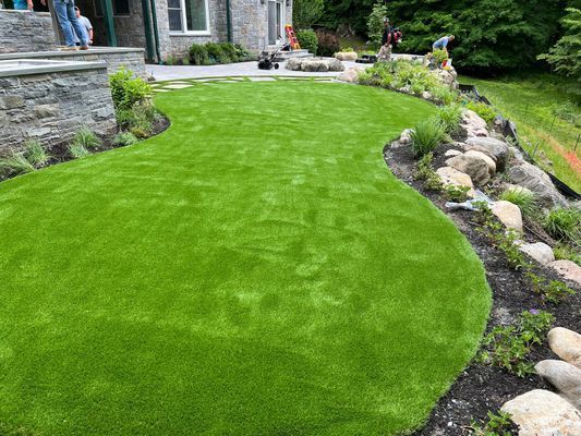 A lush, bright green lawn curves between a stone patio and a natural rock garden border near a house.