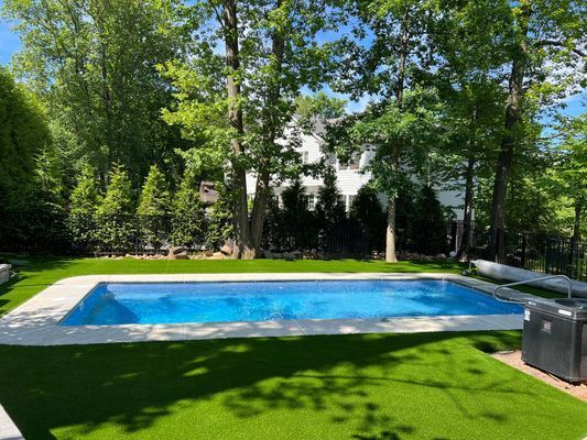 A rectangular swimming pool with bright blue water, surrounded by a light stone patio and green lawn, under leafy trees.