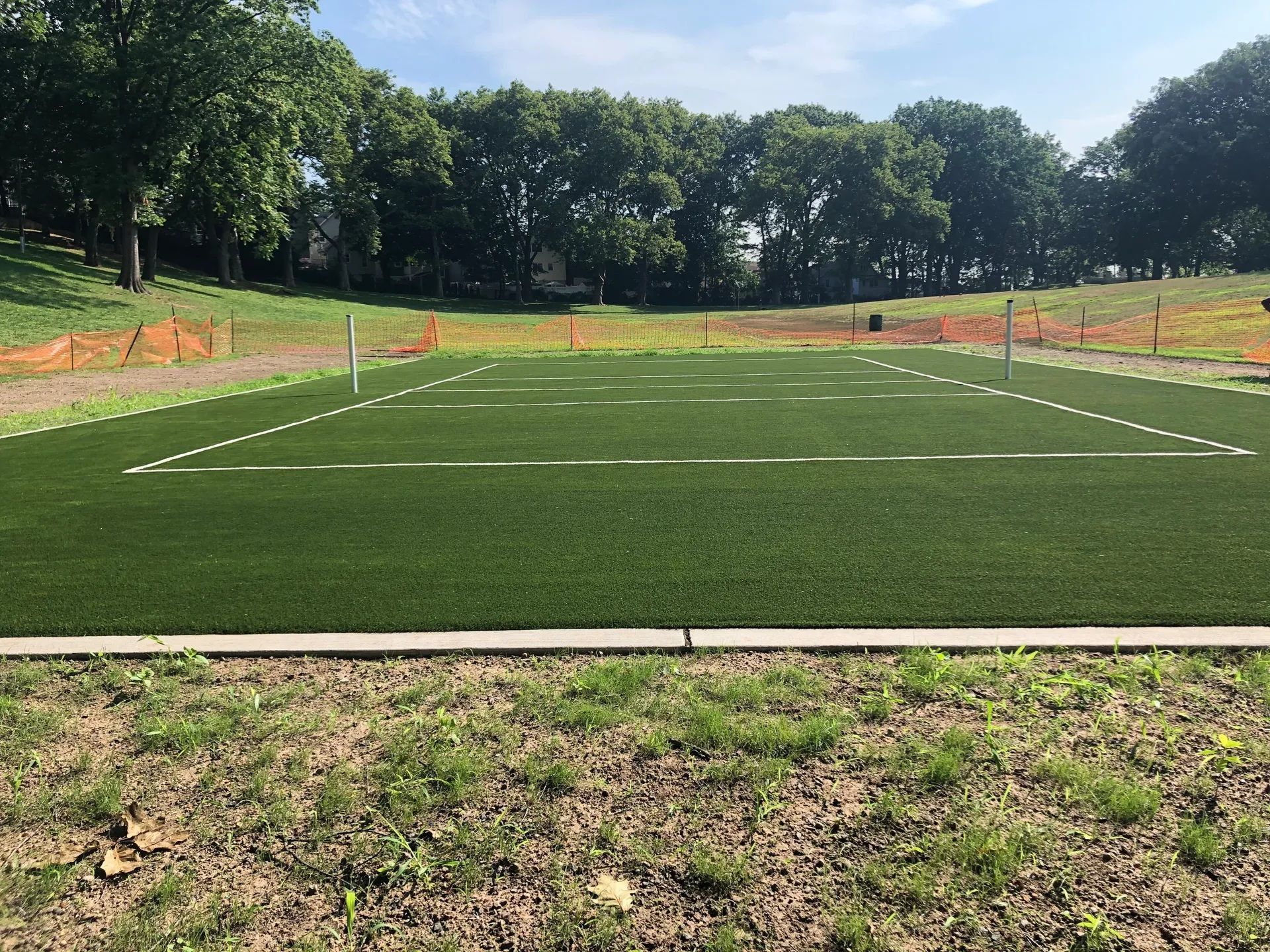 A rectangular artificial turf field with white line markings, surrounded by trees and orange construction fencing.
