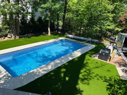 A rectangular swimming pool with bright blue water surrounded by artificial green turf and a stone border in a wooded yard.