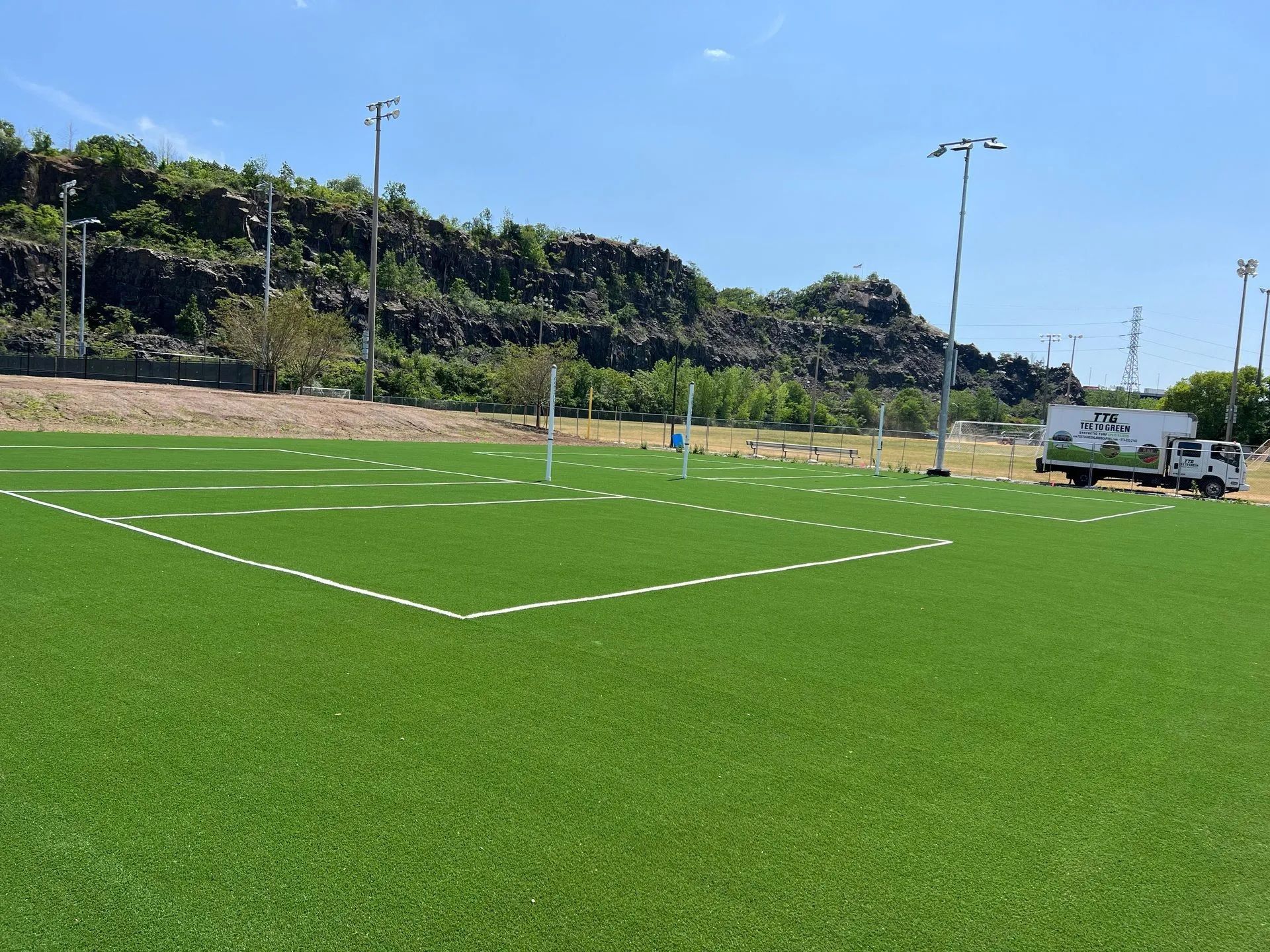 A bright green turf soccer field under a clear blue sky, bordered by a dark, rocky hillside and light poles.