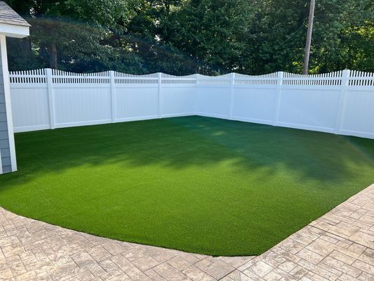 A well-manicured, vibrant green artificial lawn framed by a white vinyl fence and stone patio in a backyard setting.