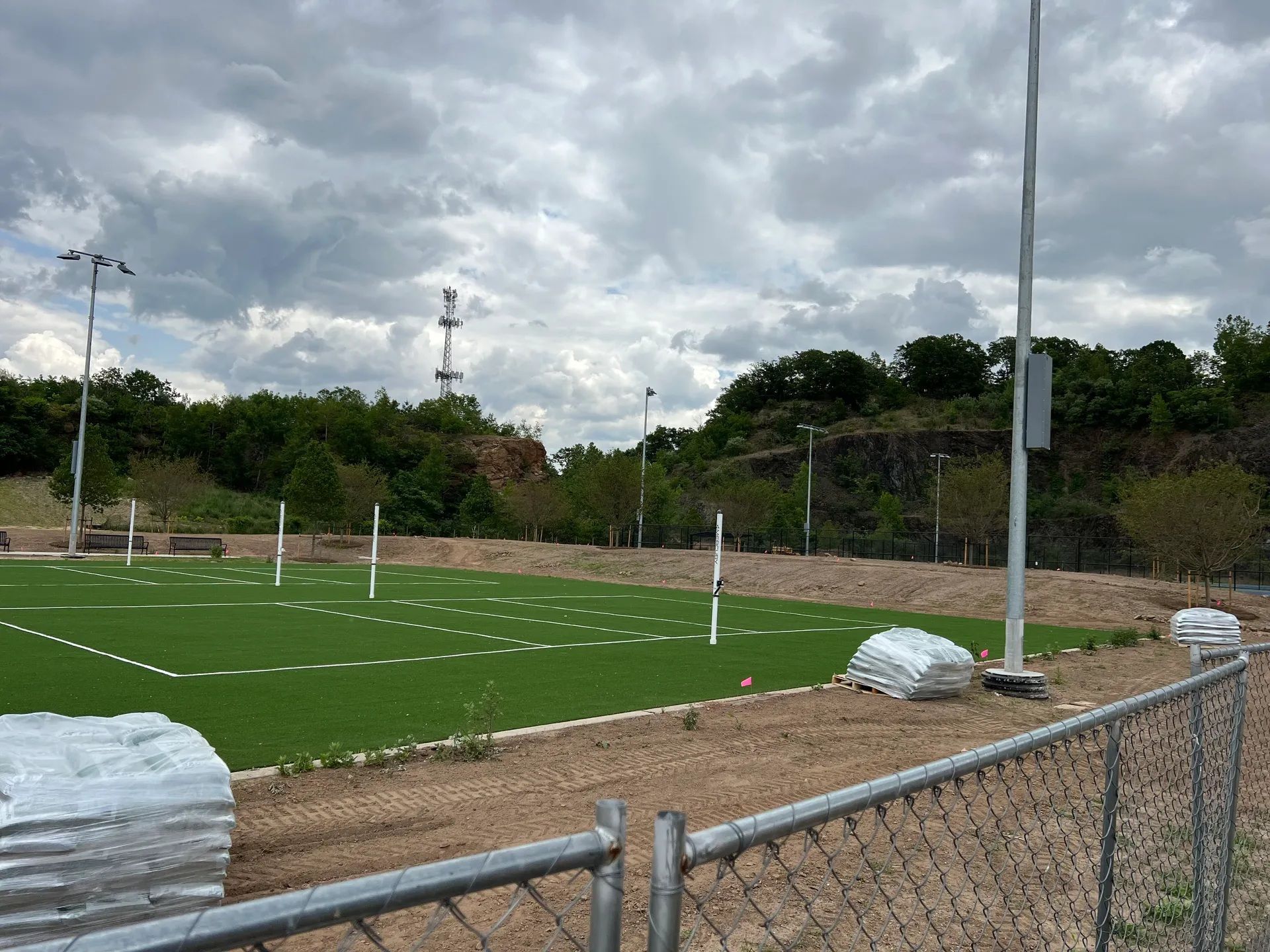 A grassy field with white boundary lines and posts sits in a construction area, enclosed by a chain-link fence.