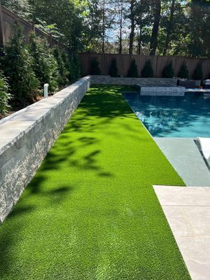 A swimming pool beside a strip of artificial green grass, lined by a light stone retaining wall and evergreen trees.
