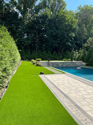 A backyard with a rectangular blue swimming pool, stone patio, green artificial lawn, and tall hedges against tall trees.