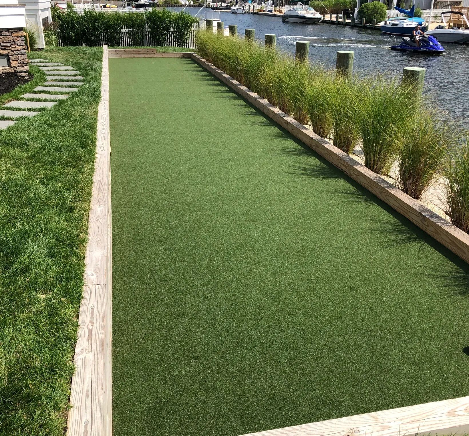 A green bocce ball court framed by wooden edges, bordered by tall grasses next to a canal with boats.