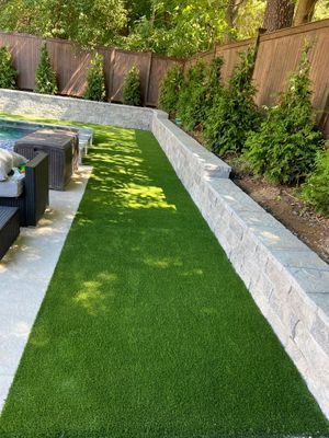 A narrow strip of green artificial turf bordering a swimming pool patio and a stone retaining wall with evergreen shrubs.