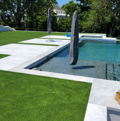 A backyard pool area with light stone pavers surrounded by vibrant green artificial turf and two closed patio umbrellas.