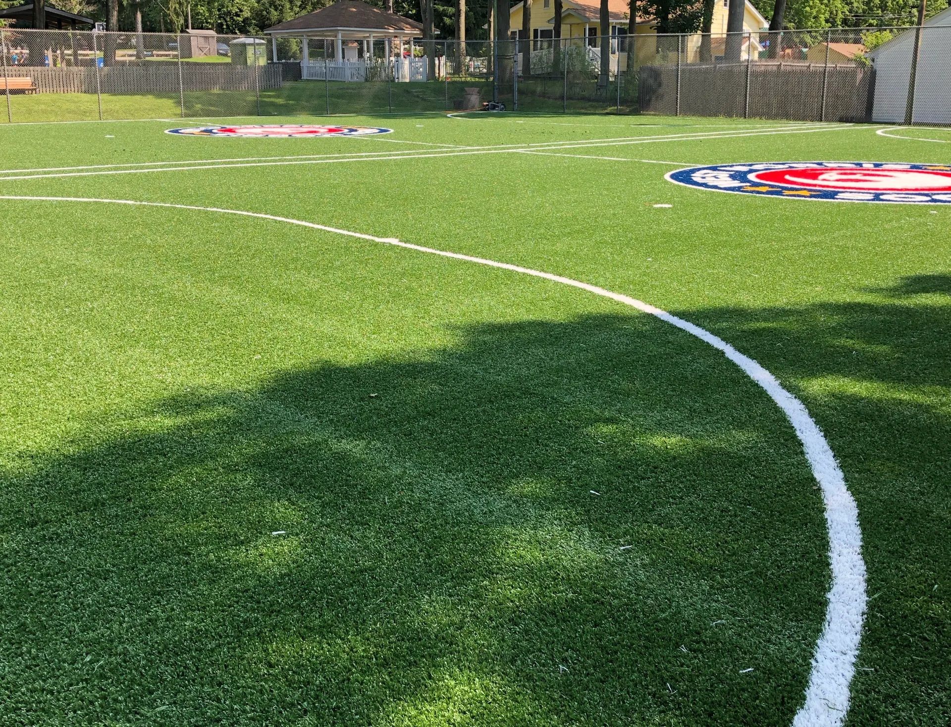 A bright green turf field with a white painted curve and two circular logos in the background.