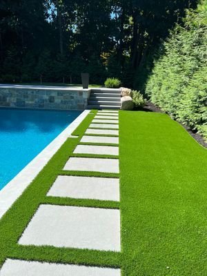 A swimming pool with a row of stone pavers leading to stone steps on a lush green lawn bordered by a tall hedge.