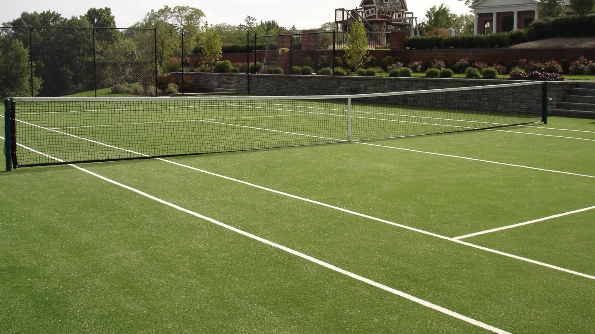 A tennis court with artificial turf surfacing and a net, situated in an outdoor area with trees and a stone retaining wall.