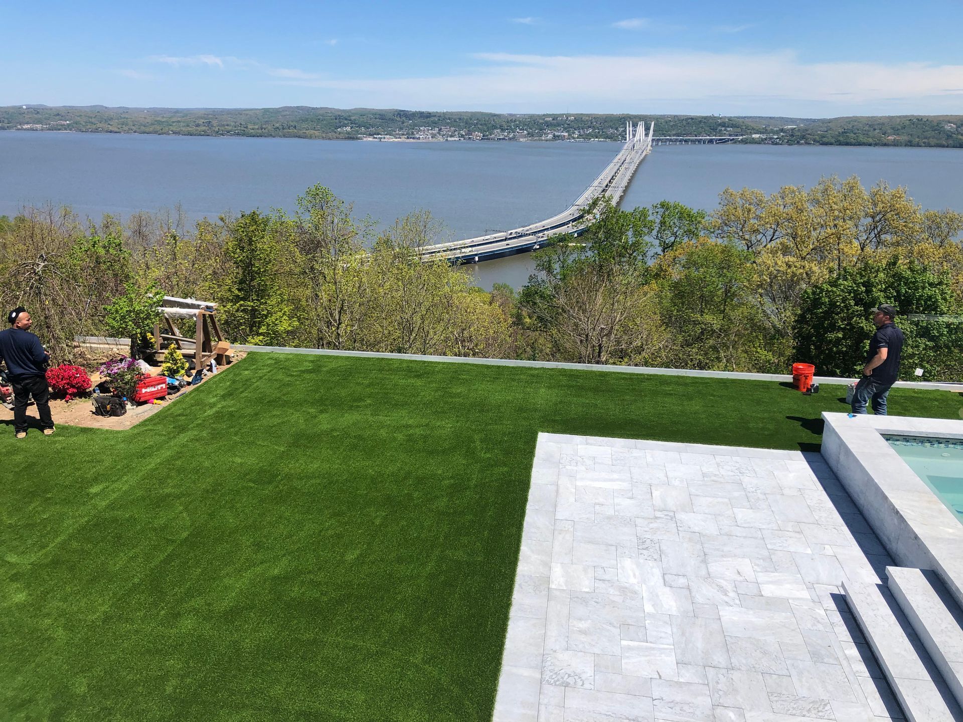 A wide view of a grassy yard with a swimming pool, two people, and a bridge stretching across a calm lake.