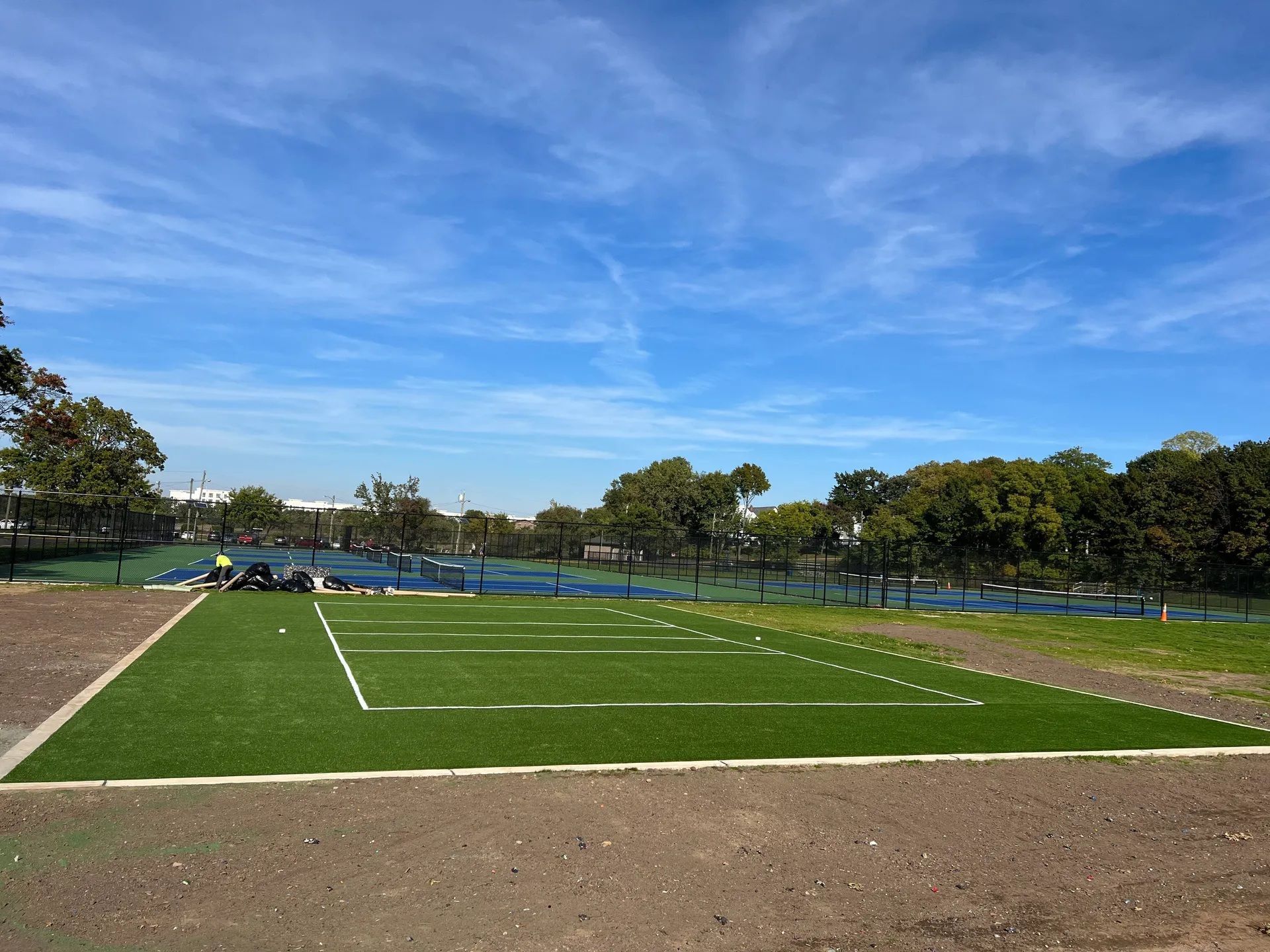 A rectangular patch of green artificial turf with white field markings, surrounded by dirt and trees under a blue sky.