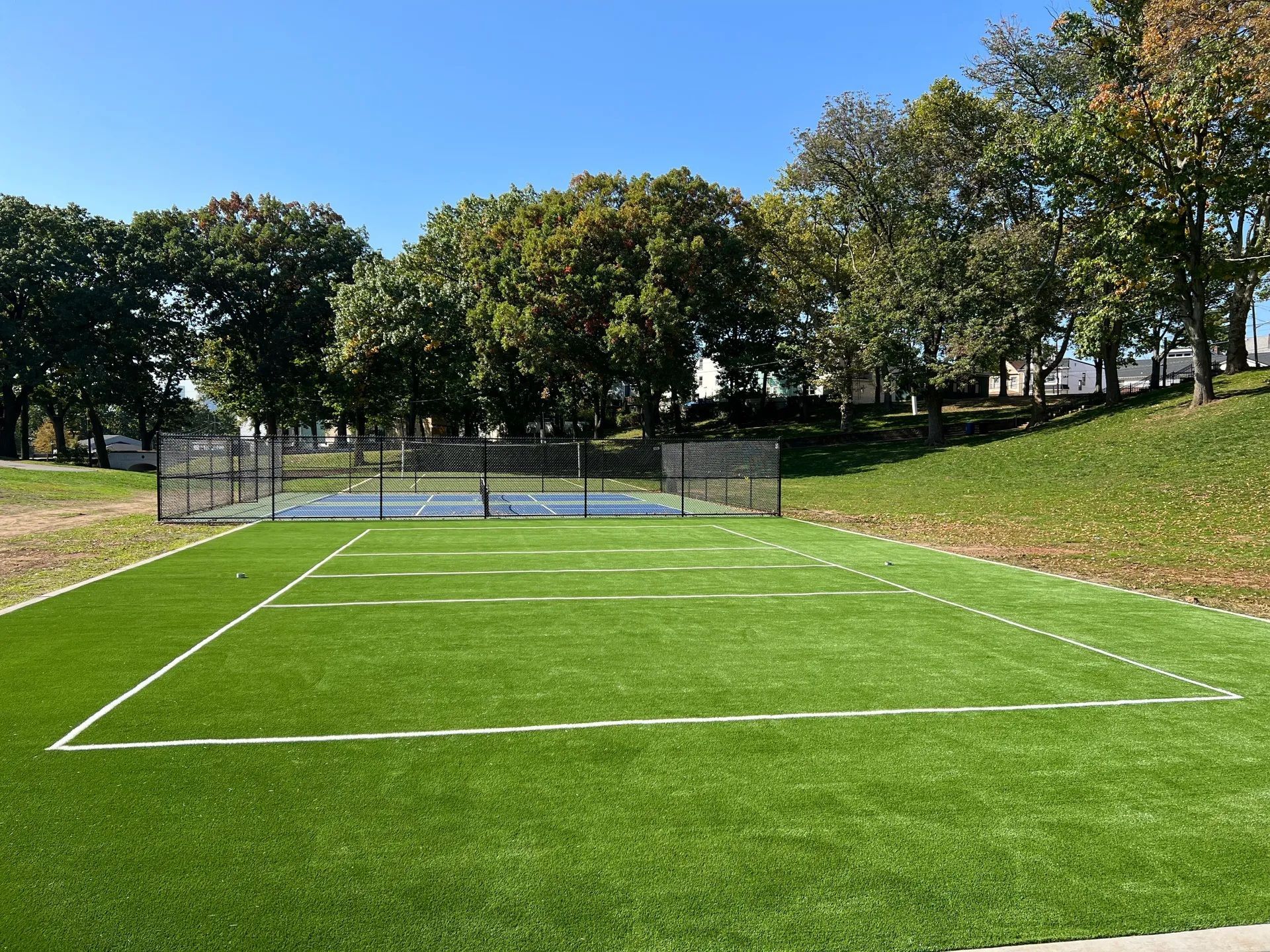 A rectangular artificial turf field with white boundary lines, set in a sunny park with trees and a fenced court nearby.