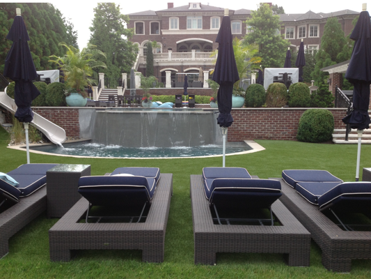A backyard scene with dark lounge chairs and umbrellas facing a stone wall with a waterfall, lawn, and a large mansion.