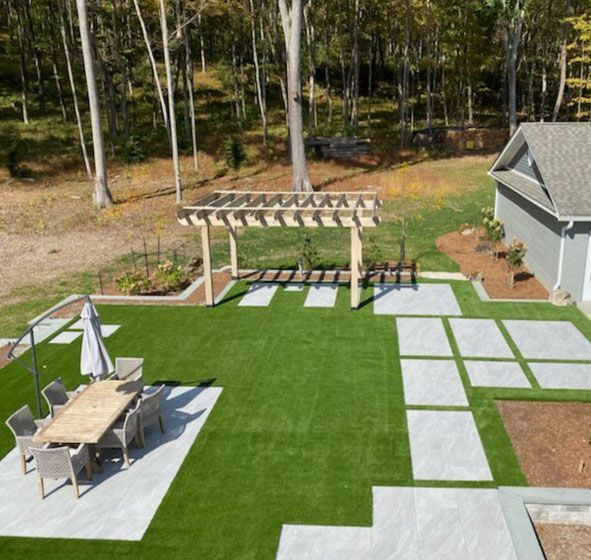 A high-angle view of a backyard with artificial turf, stone pavers, a pergola, and a dining set near a house by a woods.