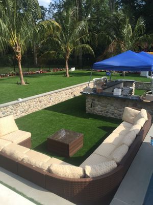 A curved wicker sofa with beige cushions surrounds a small table on a lawn near a stone bar and a blue canopy tent.