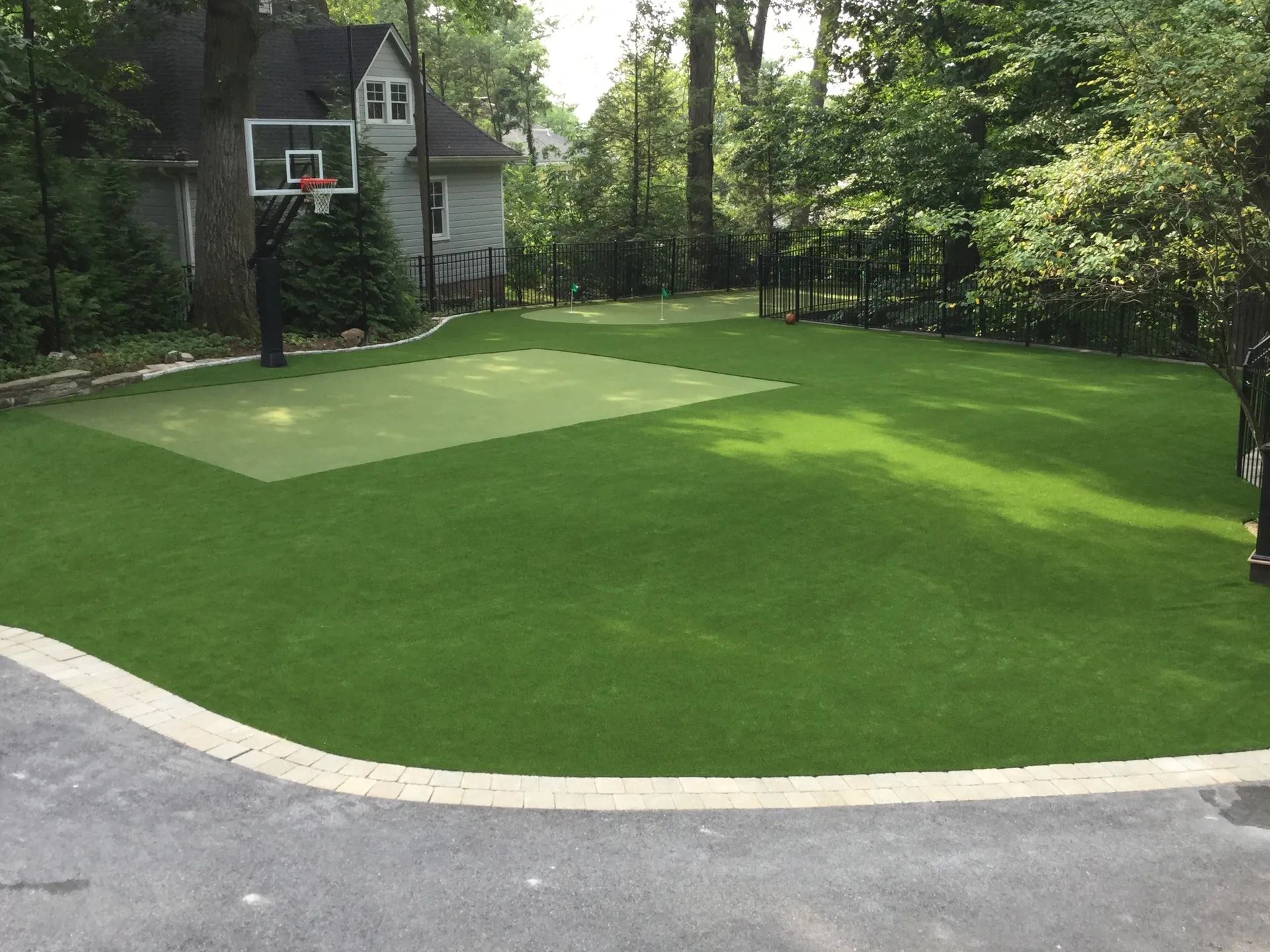 Backyard with a green artificial turf lawn, a paved basketball court area, and a basketball hoop near a house.