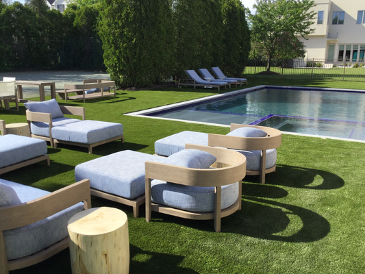 Outdoor lounge area with light blue cushions and wooden frames on grass next to a swimming pool.