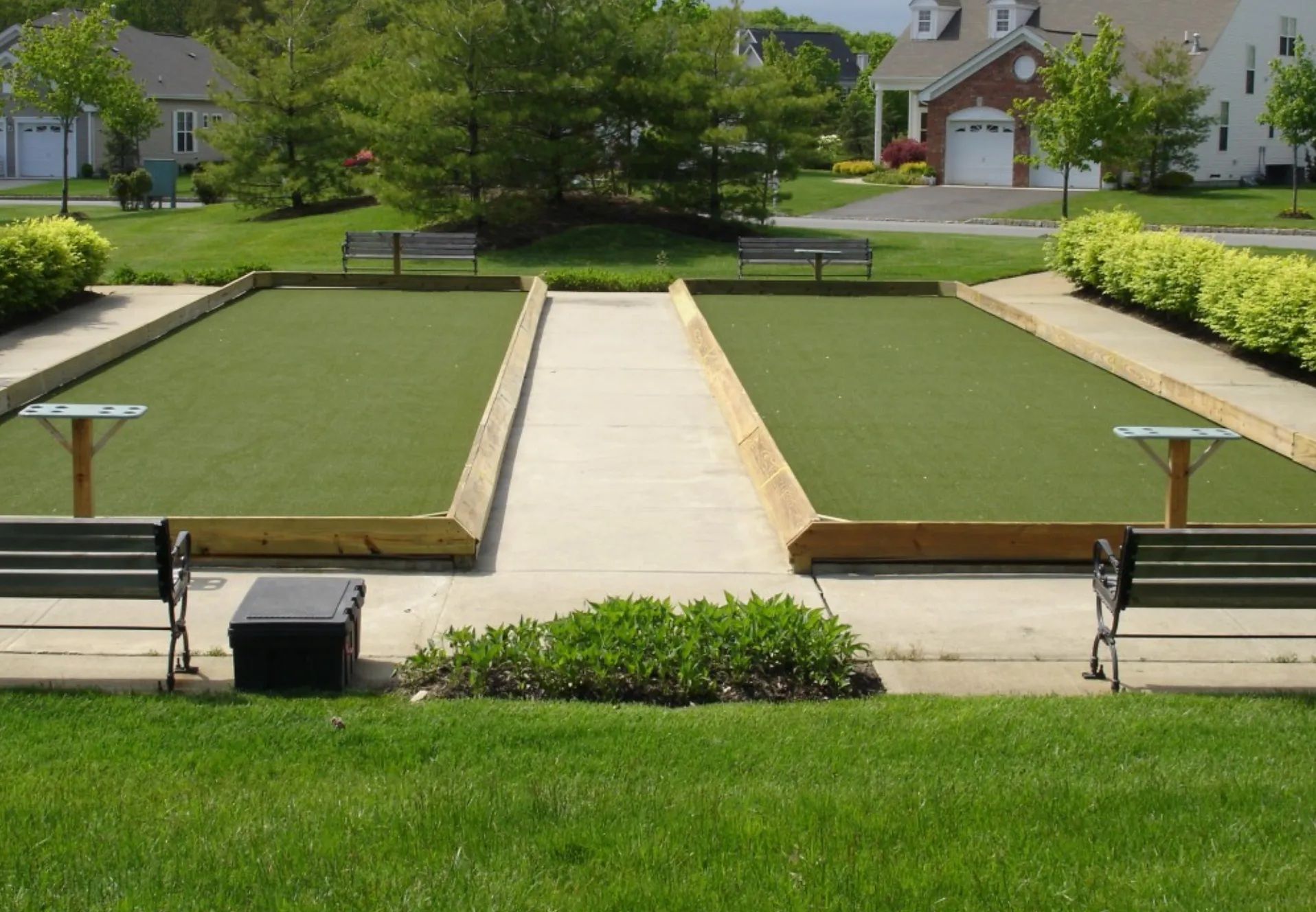 Two bocce ball courts with green artificial turf, surrounded by wooden borders, benches, and landscaping in a residential area.