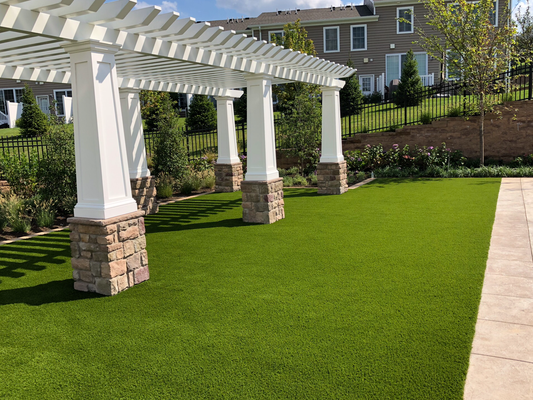A white pergola with stone-based pillars stands over a lush green artificial lawn next to a paved stone walkway.