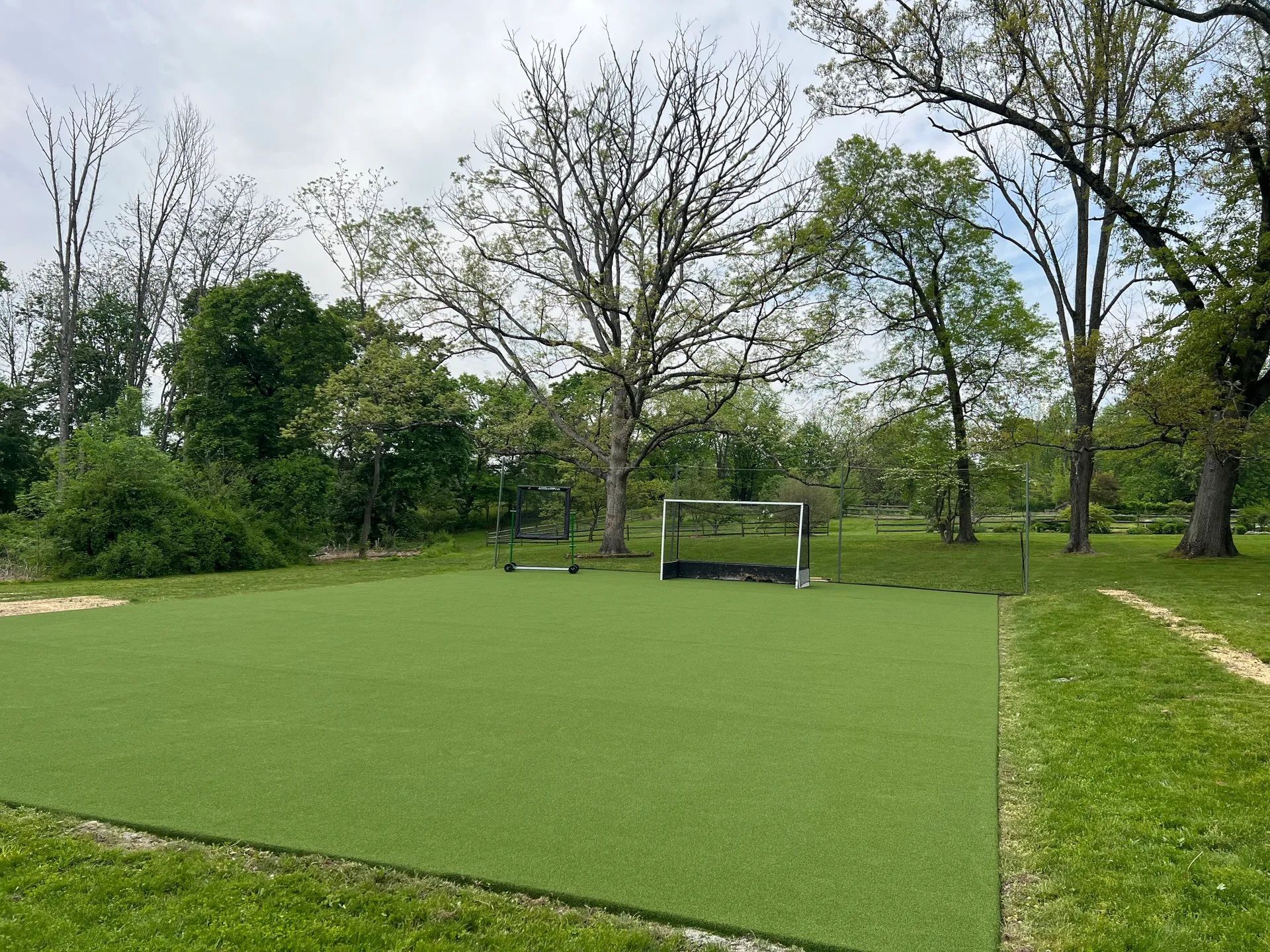 A small rectangular field with artificial green turf and a soccer goal, situated in a grassy park surrounded by trees.
