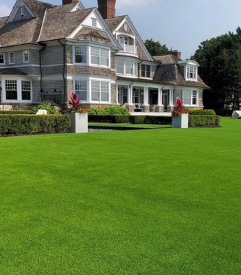 A large grey shingled mansion with a prominent green lawn, manicured hedges, and two planters with red plants.