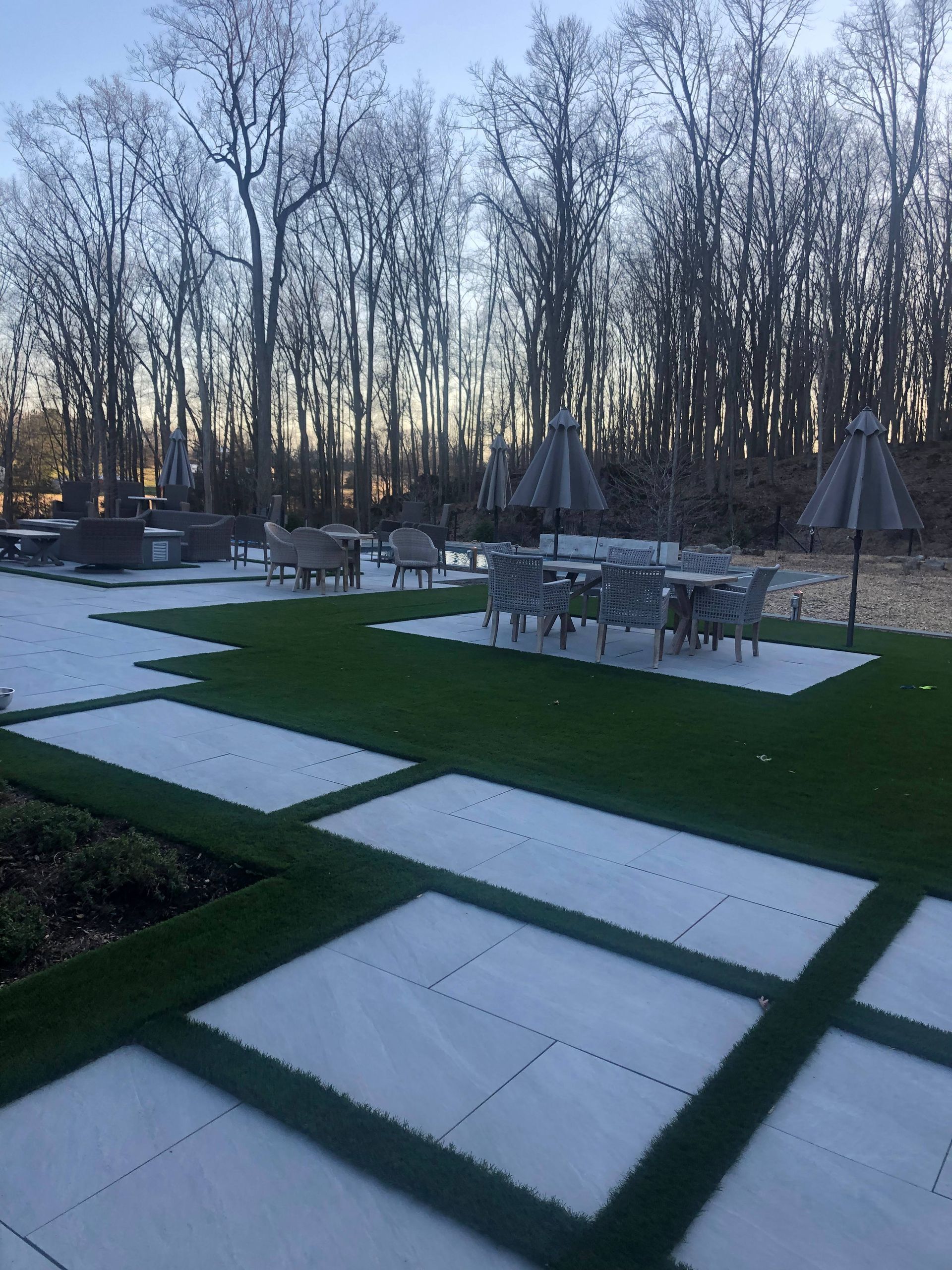 A modern outdoor patio featuring rectangular white pavers set in green artificial grass, with dining sets and trees beyond.
