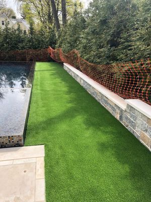 A swimming pool with artificial grass, a stone retaining wall, and an orange safety fence in a backyard.
