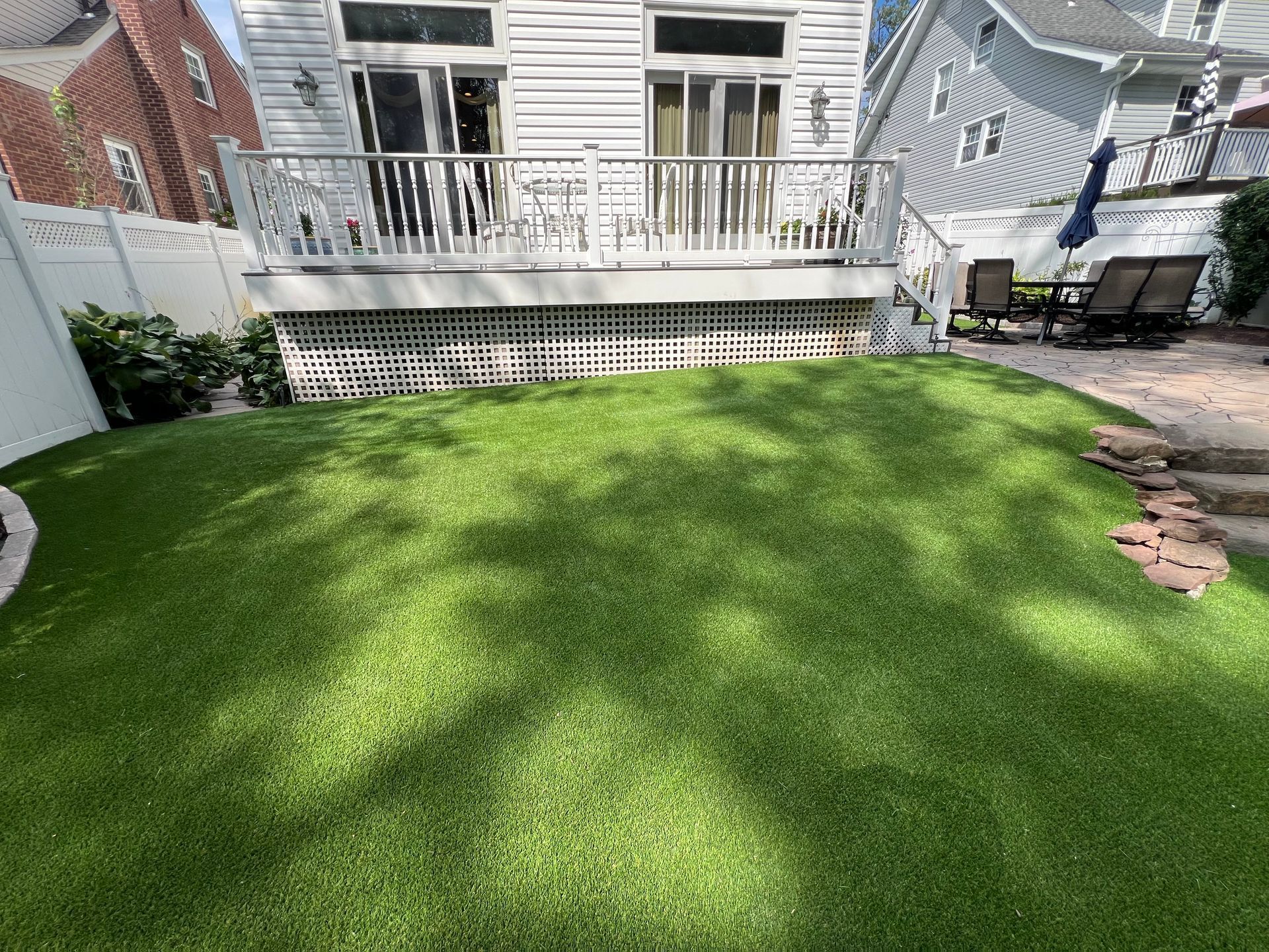 A sunny backyard featuring a green synthetic lawn, a white deck, and a stone patio area with outdoor furniture.