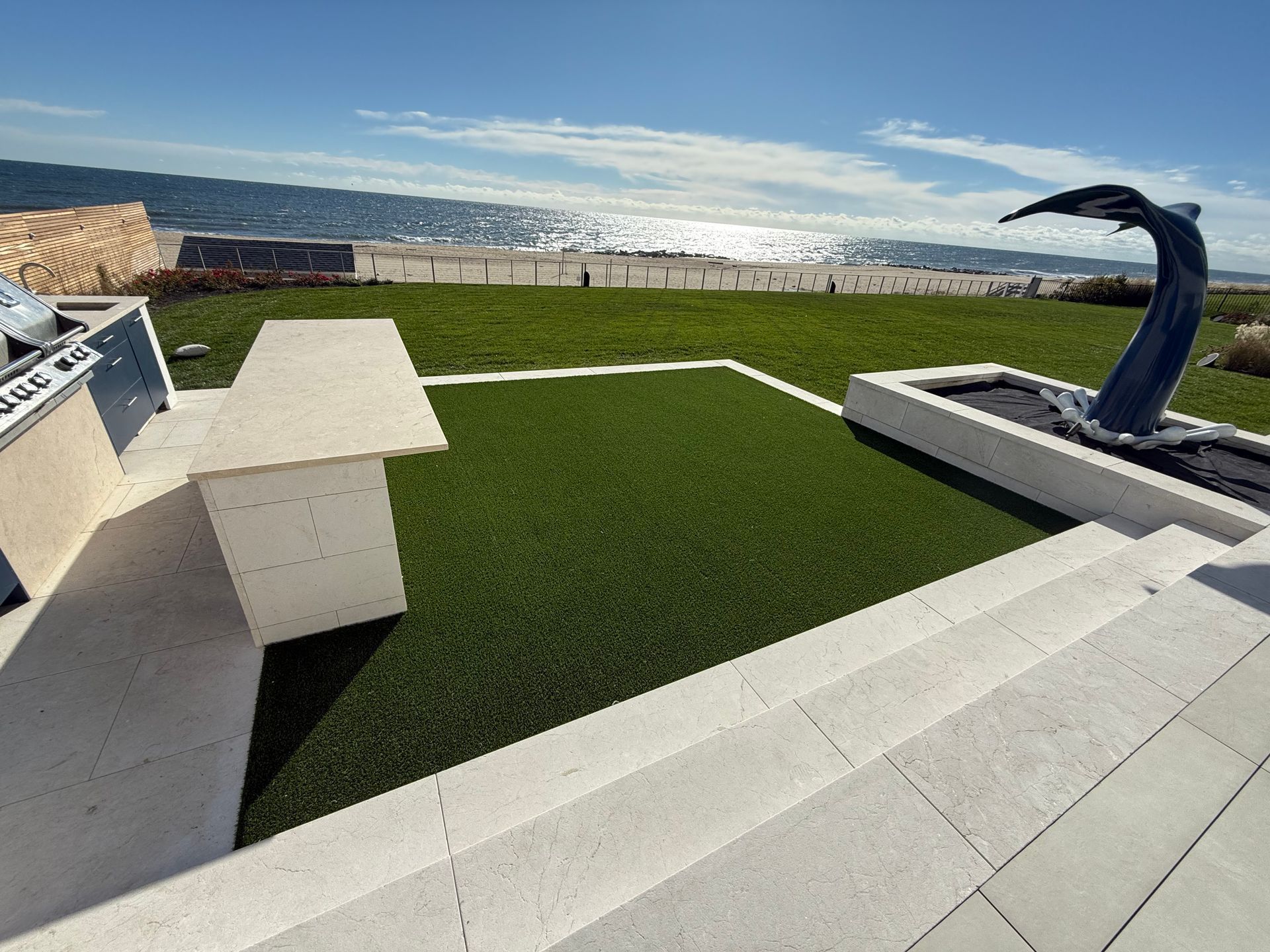 Outdoor patio with a stone bar and grill, green lawn, and a sculptural whale tail feature overlooking the ocean.