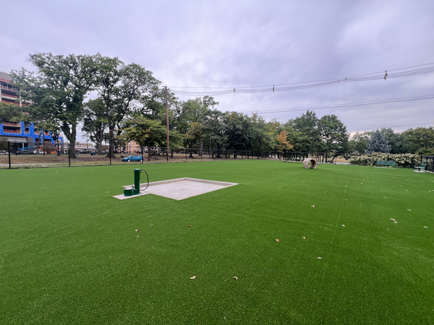 A wide outdoor park space featuring vibrant green synthetic turf, a small paved rectangular area, and perimeter trees.