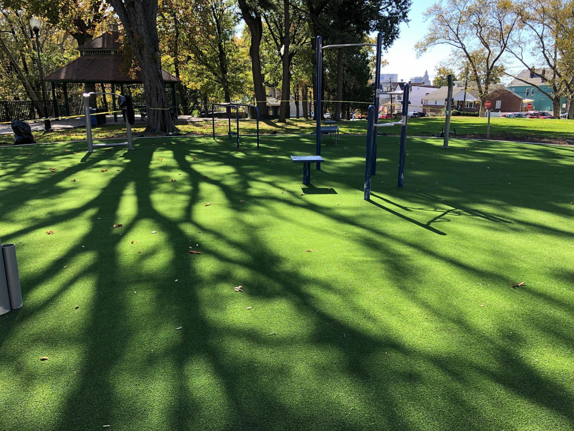 An outdoor fitness area with equipment on synthetic grass, with long tree shadows stretching across the lawn.