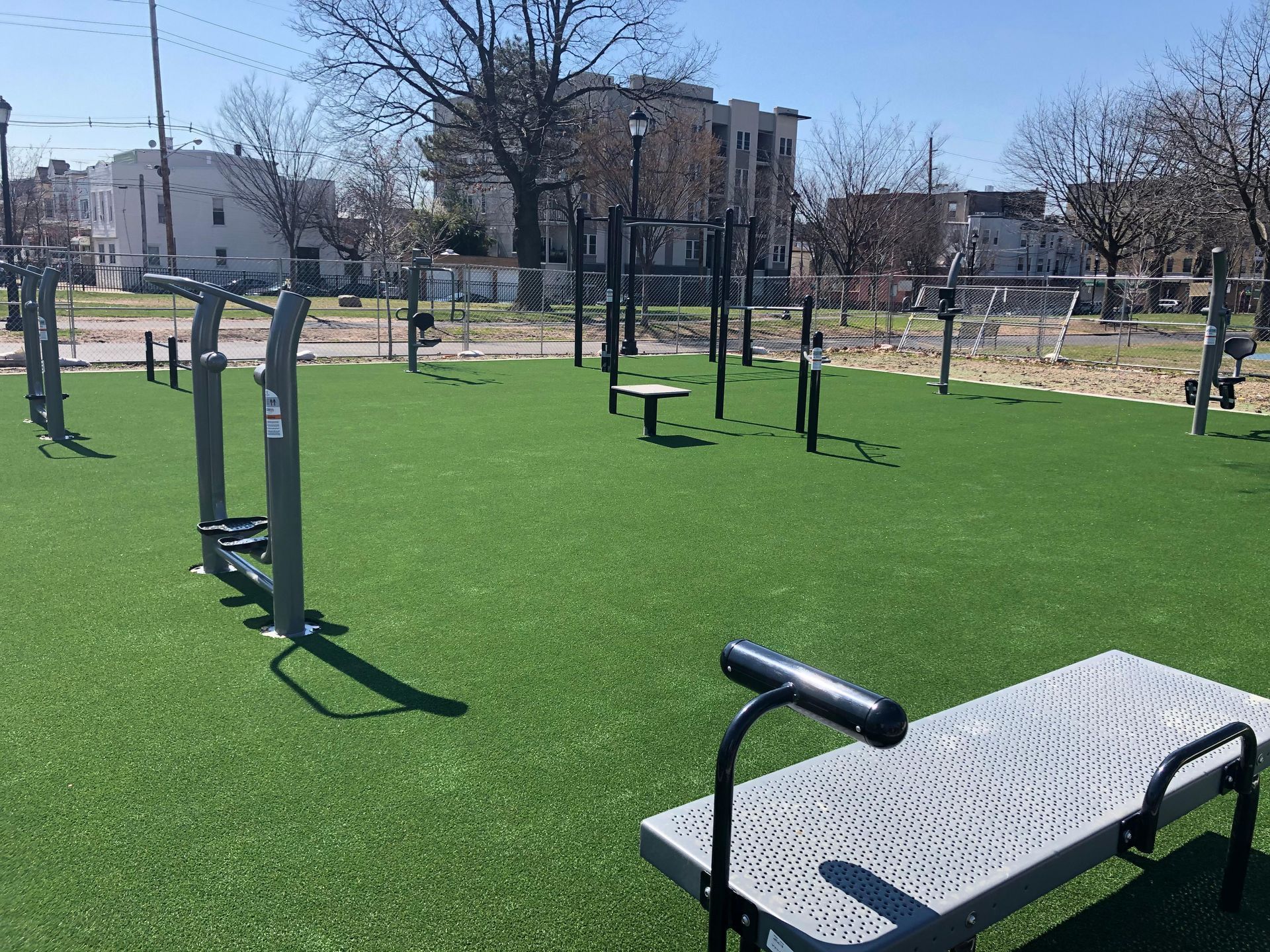 An outdoor fitness park with metal exercise equipment on a bright green turf surface under a clear blue sky.