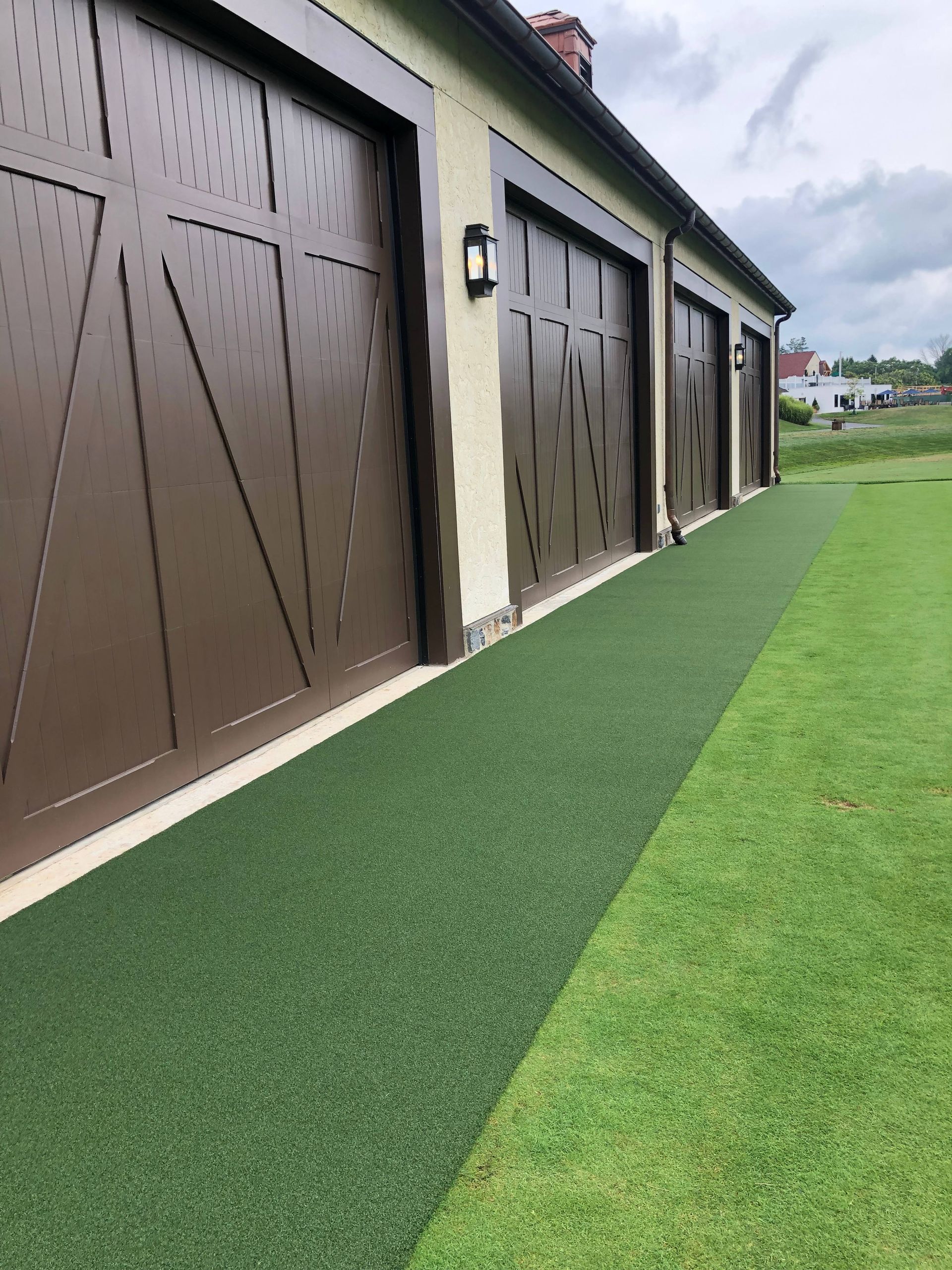 A low-angle view of brown garage doors on a light-colored building, bordered by a strip of artificial turf and green lawn.