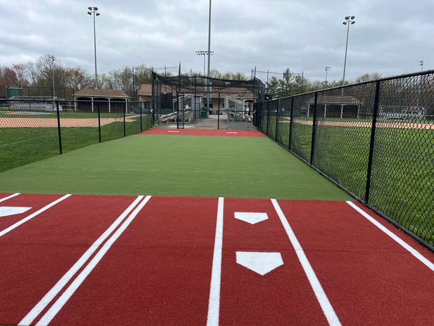 A red and green synthetic turf batting practice area with white home plate markings, enclosed by black chain-link fencing.