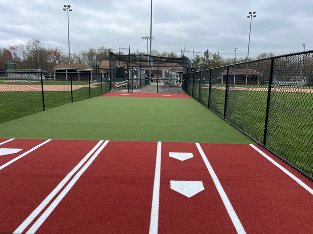 A red and green synthetic turf batting practice area with white home plate markings, enclosed by black chain-link fencing.